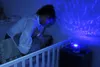 A mother is interacting with her baby in a crib, with a star projector turtle casting blue light and patterns on the ceiling. Voir les images du produit