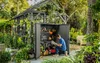 A man is organizing tools inside a gray storage shed in a garden setting. Podgląd zdjęcia produktu