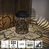 A decorative lantern with a leaf pattern is placed on a patterned surface, casting shadows. Produktbild-Vorschau