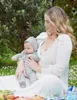 A woman is sitting on a picnic blanket outdoors, holding a baby. There is food on the blanket. Produktbild-Vorschau