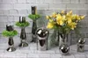 A collection of silver decorative items, including candle holders with greenery, vases with yellow flowers, and spherical ornaments, arranged on a surface against a brick wall. Produktbild-Vorschau