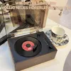 A vintage-style record player with a vinyl record on it, next to a coffee cup and saucer. Produktbild-Vorschau