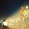 Two construction workers are discussing plans at a construction site at night, illuminated by a portable LED work light. Voir les images du produit