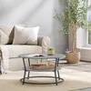 A modern coffee table with a two-tiered design is placed on a rug in front of a sofa. A small plant sits on the top tier, and a tray with items is on the lower tier. A large potted plant is to the right. Produktbild-Vorschau