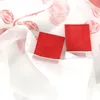 A close-up view of a white sheer curtain with a rose pattern and two red fabric squares inserted into the bottom hem. Produktbild-Vorschau