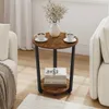 A round side table with two shelves, placed between two sofas in a living room setting. The table has a wood-grain top and black metal legs. It is decorated with a vase of flowers and two cups of coffee on the top shelf, and two books on the lower shelf. Produktbild-Vorschau