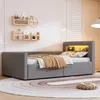 A bedroom scene featuring a gray upholstered bed with a built-in headboard shelf illuminated by warm LED lights. The bed is made with beige and brown bedding, and a round rug with concentric circles is on the floor. Produktbild-Vorschau