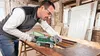 A man is sanding a wooden table with a Bosch belt sander in a workshop. Podgląd zdjęcia produktu
