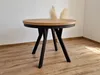 A round dining table with a wooden top and black metal legs is shown in a room with a wooden floor and a white wall. Produktbild-Vorschau