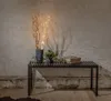 Decorative LED willow branches with lights in a vase, placed on a bench with plants and books, against a textured wall. Podgląd zdjęcia produktu