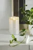 A silver LED candle with a flickering flame sits on a marble surface next to a potted plant. Podgląd zdjęcia produktu