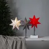 Three star-shaped lamps are displayed on a table in front of a gray wall and a Christmas tree. Podgląd zdjęcia produktu