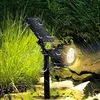 A solar-powered spotlight is shown in a garden setting during rainfall. Voir les images du produit