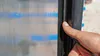 A hand is shown pressing a black sealing strip against a ribbed, translucent panel with blue markings. Produktbild-Vorschau