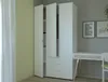 A white wardrobe with three doors and drawers, next to a desk with two framed pictures on the wall behind it. Produktbild-Vorschau