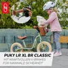 A young girl with a helmet is interacting with a green balance bike, which has a basket with stuffed animals and flowers. The bike has brown grips and a brown seat. The background shows a stone wall and greenery. Náhled obrázku produktu