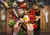 A top-down view of a person preparing a meal, with various ingredients and cooking utensils laid out on a wooden surface. Produktbild-Vorschau