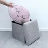 A hand is placing a pink cloud-shaped pillow with closed eyes into a gray fabric storage ottoman. A small gray hippo toy is also inside the ottoman. Náhled obrázku produktu