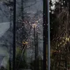 A decorative LED starburst light is displayed inside a greenhouse, with reflections of trees and the outdoors visible on the glass. Produktbild-Vorschau