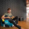 A man playing an electric guitar while sitting on a bean bag chair in a room with a brick wall. Náhľad obrázku produktu