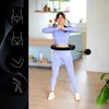 A woman is exercising with a smart hula hoop in a bright, modern room. The left side of the image features graphics illustrating body parts. Náhled obrázku produktu