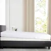 A white mattress topper is placed on a black bed frame, with a window and curtains in the background. Produktbild-Vorschau