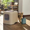 A cat stands next to a litter box and a scooper on a wooden floor, with plants and furniture visible in the background. Produktbild-Vorschau