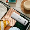 A white portable kettle is placed inside an open suitcase among folded clothes, with a straw hat visible in the background. Produktbild-Vorschau