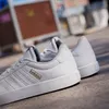 A pair of white Adidas sneakers on a textured surface, with a blurred urban background. Produktbild-Vorschau
