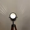 A close-up shot of a vintage-style cinema tripod floor lamp with its light on, casting shadows on a light gray wall. Anteprima dell’immagine prodotto