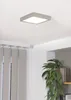 A square LED ceiling light is mounted on a white ceiling. To the left are vertical blinds, and to the right is a potted plant. Voir les images du produit