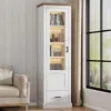 A tall, narrow display cabinet with LED lighting, filled with books and decorative items, placed next to a sofa and a window with curtains. Náhľad obrázku produktu
