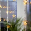 A string of star-shaped LED lights hanging in a window, with reflections of the lights and the outdoors visible. Produktbild-Vorschau
