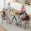 Four people are sitting around a round table, seemingly in a modern dining area or kitchen. The table has a wood-grain top and black legs. There are cups and a bowl of oranges on the table. The background shows white cabinets, some appliances, and a floor  Produktbild-Vorschau