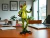 A frog figurine is placed on a wooden desk next to a laptop and a notebook. Produktbild-Vorschau