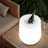 An illuminated ice bucket with a bottle of champagne sits on a woven table next to a wicker chair, with greenery in the background. Náhled obrázku produktu