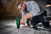 A man is kneeling on a wooden floor, using a cordless drill to screw into the floorboards. The scene appears to be in an attic or workshop, with hay bales and a ladder visible in the background. Produktbild-Vorschau