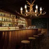 A dimly lit bar interior with a prominent antler chandelier, shelves stocked with bottles, and a wooden bar counter with stools. Podgląd zdjęcia produktu