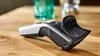 A hair clipper is placed on a wooden surface, with blurred glasses in the background. Anteprima dell’immagine prodotto