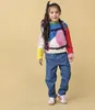 A young girl wearing a colorful sweater and jeans, with a backpack on, stands against a plain yellow background. Produktbild-Vorschau