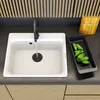 A white kitchen sink with a black faucet running water into it, next to a black colander filled with cucumbers. Podgląd zdjęcia produktu