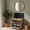 A shoe cabinet with a flip-top storage compartment and multiple shelves filled with shoes, placed next to a large potted plant and a round mirror on a green wall. Náhľad obrázku produktu