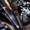 A close-up view of folded fabric with a dark, abstract floral or feather-like pattern in black, gray, white, and orange. Produktbild-Vorschau
