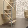 A modern spiral staircase with wooden steps and a metal railing is shown in a room with a stone wall and a chair. Produktbild-Vorschau