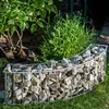 A curved gabion planter filled with stones, set in a garden with green plants and grass. Produktbild-Vorschau