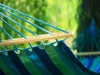 A close-up view of a striped hammock made of fabric and supported by a wooden spreader bar and blue ropes, set against a blurred green background of foliage. Produktbild-Vorschau