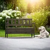 A dark brown wooden garden bench with a storage compartment is placed on a paved patio next to a lush green garden with white flowers. A golden retriever dog is lying down on the patio in front of the bench. Voir les images du produit
