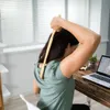 A woman is sitting at a desk and using a back scratcher on her upper back. A laptop is on the desk. Produktbild-Vorschau