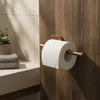 A toilet paper holder with a roll of toilet paper mounted on a wooden wall, with a shelf and towels in the background. Produktbild-Vorschau