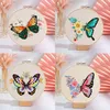 A collage of four embroidered hoop art pieces featuring butterflies and flowers, displayed on a white surface with decorative pink fans and vases in the background. Produktbild-Vorschau